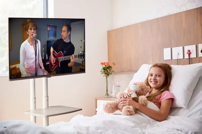 Young patient in hospital bed watching live musicians perform via video screen during virtual Harmony & Healing performance