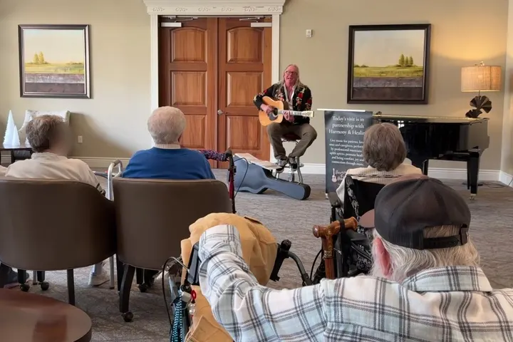 A Harmony & Healing artist plays acoustic guitar for residents gathered in a memory care day room