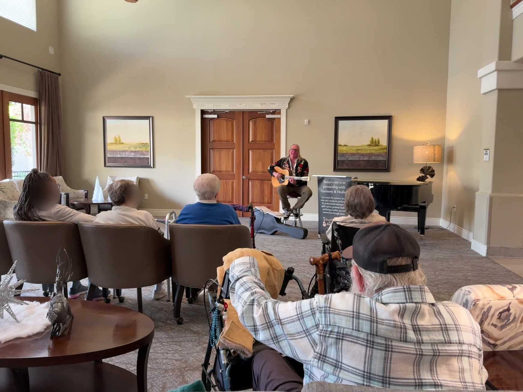 A guitarist sings by the piano while patients sit in armchairs around a bright memory care common room.