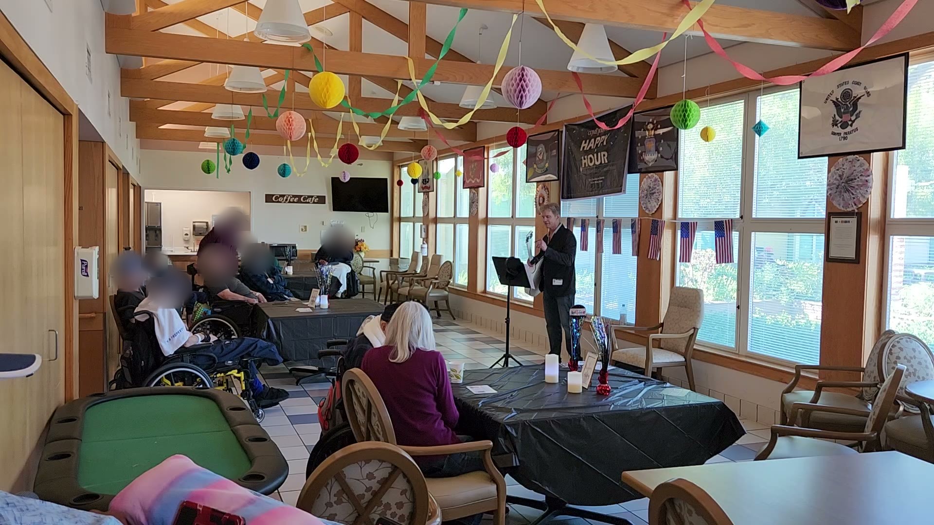 Veterans gather at tables in the Martinez VA day room, decorated with streamers, as a musician plays at a mic stand by the windows.